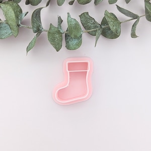 May include: A pink plastic Christmas stocking cookie cutter. The cutter is on a white background with a sprig of eucalyptus leaves above it.