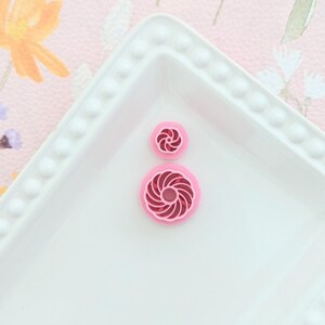 May include: Two pink plastic cookie cutters in the shape of a swirl. The cutters are on a white plate with a beaded edge.