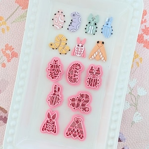 May include: A selection of colourful insect-shaped embellishments in various designs, including beetles, caterpillars, and moths. The embellishments are in shades of pink, yellow, blue, and purple, arranged on a white tray.