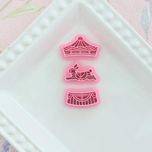 May include: Three pink cookie cutters shaped as a crown, a rabbit, and a circus tent. They are on a white, rectangular plate with a beaded edge. The background features a floral pattern.