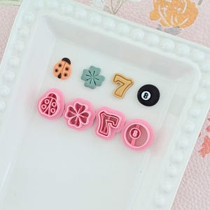 May include: A selection of colourful polymer clay charms displayed in a white tray. The charms include a ladybug, a four-leaf clover, the number 7, a black 8-ball, and pink cookie cutters shaped like a ladybug, a flower, the number 7, and an 8-ball.