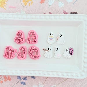 Op de afbeelding: Een witte rechthoekige schaal bevat roze koekjessnijders en spookvormige versieringen. De koekjessnijders hebben de vorm van geesten en andere figuren. De versieringen zijn witte geesten met verschillende ontwerpen.