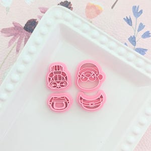 Puede incluir: Cuatro cortadores de galletas de plástico rosa en forma de mujer con un moño, un Papá Noel, un torso de mujer y un torso de Papá Noel.