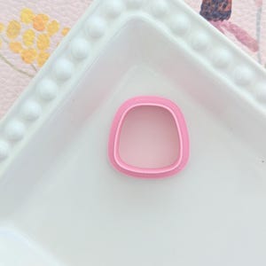 May include: A pink, square-shaped cookie cutter with rounded corners sits on a white, square plate with a beaded edge. The background is a pink floral pattern.