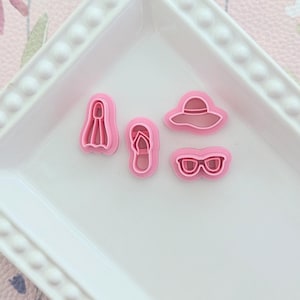 Könnte beinhalten: Vier rosa Ausstecher in Form von Sommerartikeln: Flossen, Flip-Flops, Sonnenhut und Sonnenbrille. Sie sind auf einem weißen, quadratischen Teller mit Perlenrand angeordnet. Der Hintergrund ist ein rosa Blumenmuster.