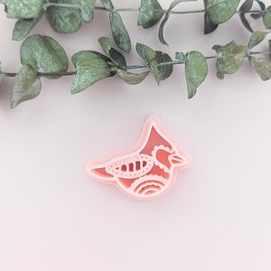 Op de afbeelding: Een roze plastic koekjesvormpje in de vorm van een kardinaal. De vogel heeft een gedetailleerd ontwerp met een kuif op zijn kop.