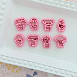 May include: A collection of pink mushroom-shaped cookie cutters in various designs. The cutters are arranged on a white rectangular tray with a decorative edge, set against a floral background. Ideal for baking themed treats.