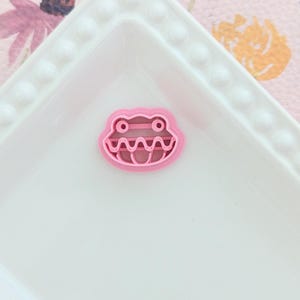 May include: A pink frog-shaped cookie cutter on a white rectangular plate with a beaded edge. The frog cutter has a detailed outline with eyes and a wide, toothy grin. The background features a floral pattern.
