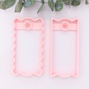 May include: Two pink plastic cookie cutters shaped like rectangular tags with scalloped edges and a hole at the top.
