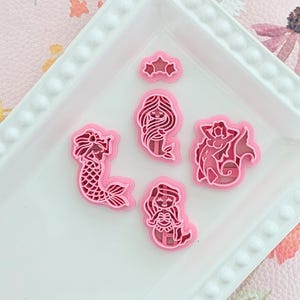 May include: Pink mermaid-shaped cookie cutters and a star-shaped cutter on a white tray. The cutters are made of plastic and are in various mermaid poses. The tray sits on a pink floral patterned surface.