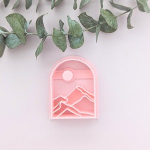 May include: Pink plastic cookie cutter shaped like a mountain range with a sun in the sky.