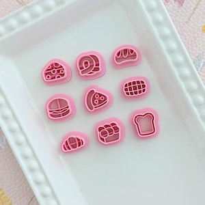 Puede incluir: Una colección de cortadores de galletas rosas con forma de alimentos, que incluyen una rebanada de queso, un caracol y una rebanada de pan, están dispuestos en un plato rectangular blanco. El plato está sobre un fondo con estampado floral.