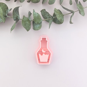 May include: A pink, potion-shaped cookie cutter with a moon and crown design. The cutter is on a white surface, with green eucalyptus leaves in the background.