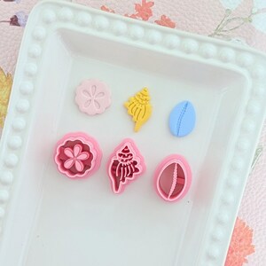 May include: A white rectangular tray holds six colorful cookie cutters in the shapes of seashells and flowers. The cutters are pink, yellow, and blue. The tray has a decorative beaded edge.