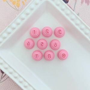 May include: Pink plastic number stamps, 1 through 8 and 0, on a white plate with a beaded edge.