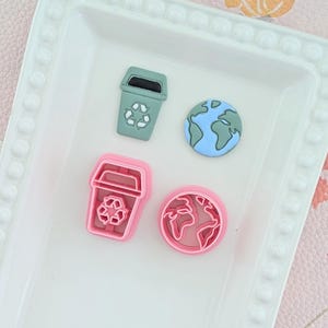 May include: A set of four items including a sage green recycling bin charm, a circular earth charm with blue and green details, a pink recycling bin cookie cutter, and a pink circular earth cookie cutter, all arranged on a white tray.