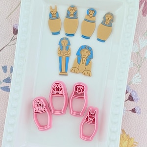 May include: A collection of Egyptian-themed craft supplies. The image features several tan and blue sarcophagus and pharaoh figures, along with pink sarcophagus-shaped cookie cutters. The items are arranged on a white tray with a decorative border.