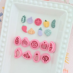 May include: A white rectangular tray displays a collection of colourful vegetable-shaped cookie cutters. The cutters are in shades of pink, red, yellow, green, and purple. The tray has a beaded edge. The vegetables include a tomato, pepper, and onion.