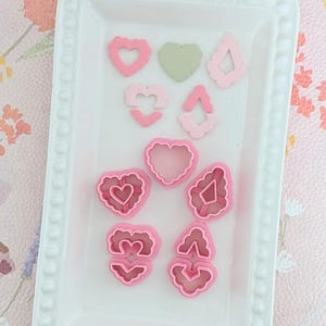 Può includere: Una collezione di formine per biscotti a forma di cuore rosa e verde in vari disegni, tra cui cuori pieni, cuori delineati e cuori con dettagli interni. Le formine sono disposte su un vassoio rettangolare bianco.