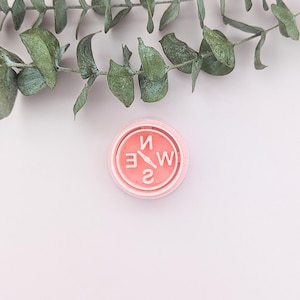 Può includere: Un timbro a forma di bussola rosa con le lettere N, W, E e S in bianco. Il timbro è su uno sfondo bianco con un rametto di foglie di eucalipto.