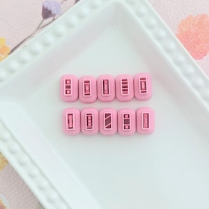 Puede incluir: Un conjunto de 10 punzones de artesanía de plástico rosa con diferentes diseños, que incluyen rayas, puntos y formas geométricas. Los punzones están dispuestos en dos filas sobre un plato blanco.