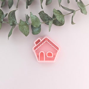 May include: Pink plastic gingerbread house cookie cutter. The cutter has a detailed design with a chimney, windows, and a scalloped roof.