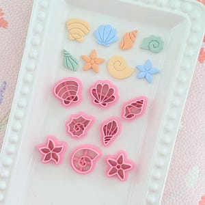 Op de afbeelding: Een verzameling schelpvormige knutselmaterialen in verschillende kleuren, waaronder roze, blauw, oranje en geel. De collectie bevat zeesterren en schelpontwerpen, gerangschikt op een wit dienblad met een parelrand. Voor knutselen of decoratie.