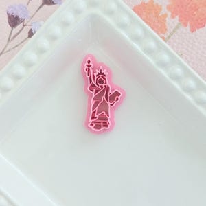 Può includere: Un ciondolo rosa e rosso della Statua della Libertà. Il ciondolo è un design a strati con un contorno rosa chiaro e un interno rosso più scuro. È posto su un piatto rettangolare bianco con bordo perlato.