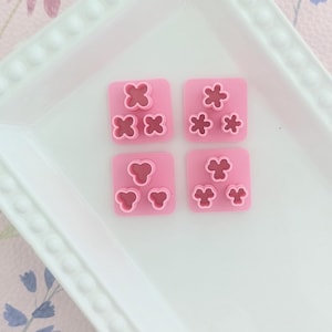 Peut inclure: Quatre emporte-pièces en plastique rose avec différentes formes de fleurs. Les emporte-pièces sont disposés en deux rangées de deux sur une assiette blanche.