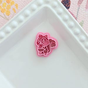 May include: A pink and red squirrel-shaped charm on a white square dish. The charm has a detailed outline and is holding a nut. The dish has a beaded edge and sits on a pink floral patterned surface.