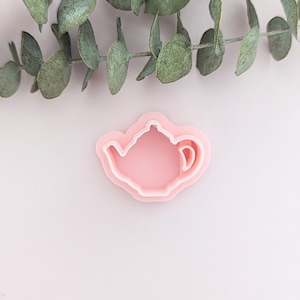 May include: A pink teapot-shaped cookie cutter with a white outline on a light pink surface. Eucalyptus leaves are arranged at the top. This kitchen tool is for baking biscuits or cookies.