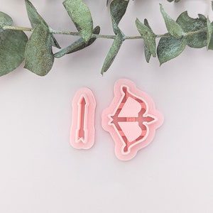 Puede incluir: Cortadores de galletas de plástico rosa en forma de arco y flecha. El arco y la flecha son cortadores separados.