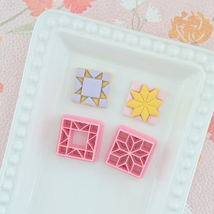 Könnte beinhalten: Zwei quadratische Ausstechformen aus rosa Kunststoff mit komplexen geometrischen Mustern und zwei passende quadratische Formen aus Ton oder Fondant in Helllila und Gelb, angeordnet auf einem weißen rechteckigen Tablett mit Perlenrand.
