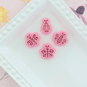Puede incluir: Un plato rectangular blanco con borde de cuentas contiene cuatro cortadores de galletas rosas con forma de insectos. Los cortadores incluyen una mariposa, una libélula, una abeja y una mariquita. El fondo es un estampado floral.