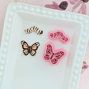 May include: A selection of insect-themed embellishments, including two butterflies and two caterpillars, presented on a white rectangular tray. The butterflies are in orange, black, and pink, while the caterpillars are in shades of pink and yellow.