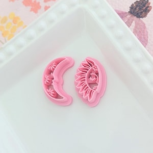 Op de afbeelding: Twee roze plastic koekjesvormen, een in de vorm van een halve maan met bloemen en de andere in de vorm van een zon met een gezicht en bloemen.