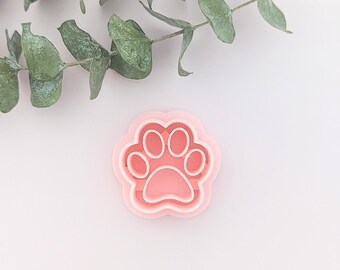 May include: A pink plastic paw print cookie cutter. The cutter has a detailed design with four toes and a pad.