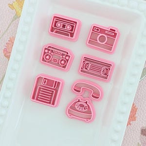May include: A selection of pink plastic cookie cutters shaped like a cassette tape, camera, boombox, VHS tape, floppy disk, and a rotary phone. The cutters are in a white rectangular tray.