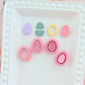 Peut inclure: Une collection d'ornements en forme d'œufs de Pâques colorés dans diverses nuances de rose, jaune, vert et violet. Les œufs sont disposés sur un plateau blanc décoratif, idéal pour l'artisanat ou la décoration saisonnière.