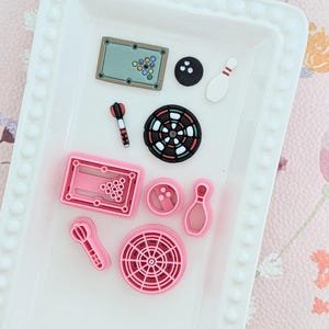 Op de afbeelding: Een verzameling miniatuur spelthema bedels, waaronder een pooltafel, bowlingpin, dart, dartbord en bowlingbal. De bedels zijn in verschillende kleuren, waaronder roze, zwart, wit en groen. De bedels zijn gerangschikt op een wit dienblad.