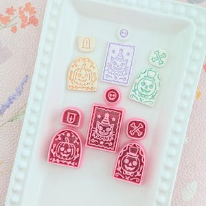 Peut inclure: Un ensemble d'emporte-pièces en plastique rose et leurs empreintes en pâte claire. Le lot comprend des motifs de citrouille d'Halloween, de chat avec un chapeau de fête, de crâne et d'os croisés, disposés sur un plateau rectangulaire blanc.
