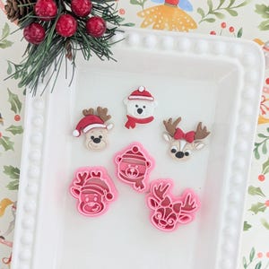 Könnte beinhalten: Ein weißes rechteckiges Tablett enthält sechs weihnachtliche Verzierungen. Die Verzierungen umfassen ein Rentier, einen Eisbären und ein Reh, alle mit festlichen Hüten und Schals. Es gibt auch rosa Ausstecher in Form derselben Charaktere.