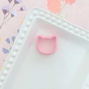 May include: A pink plastic cat-shaped cookie cutter on a white plate with a floral pattern.