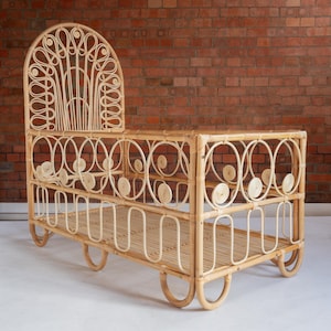 May include: A natural woven rattan cot with a unique arched headboard featuring a decorative pattern of circles and swirls. The cot has a woven rattan base and legs.