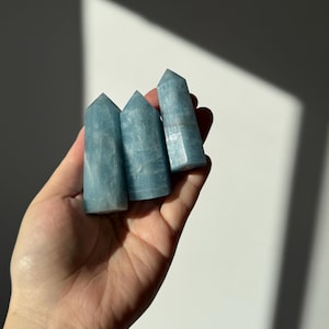 Puede incluir: Tres torres de cristal de aguamarina azul claro con puntas. Los cristales se sostienen en una mano, con una pared blanca de fondo. Los cristales tienen un aspecto translúcido y son de color azul pálido.