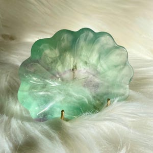 May include: A green and purple fluorite crystal bowl with a scalloped edge. The bowl is supported by two small gold-coloured metal posts and sits on a white, fluffy surface. The bowl's colours range from light green to purple.