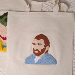May include: A white canvas tote bag with a colorful embroidered portrait of Vincent van Gogh. The portrait is in a blue shirt with a brown beard and mustache.