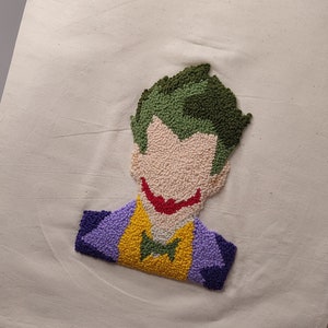 May include: A white canvas tote bag with a green, yellow, purple, and red embroidered image of the Joker from Batman.