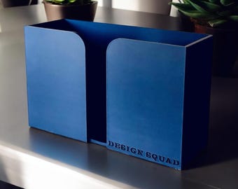 May include: A blue metal organizer with a rectangular shape and curved cutouts. The text "DESIGN SQUAD" is embossed on the side. The organizer is sitting on a metallic surface, with potted plants in the background.