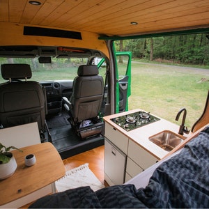Puede incluir: El interior de una furgoneta convertida con una mesa pequeña, una planta y una cama. La furgoneta tiene una zona de cocina con un fregadero, una estufa y armarios. La puerta trasera está abierta, revelando una vista de un área verde.